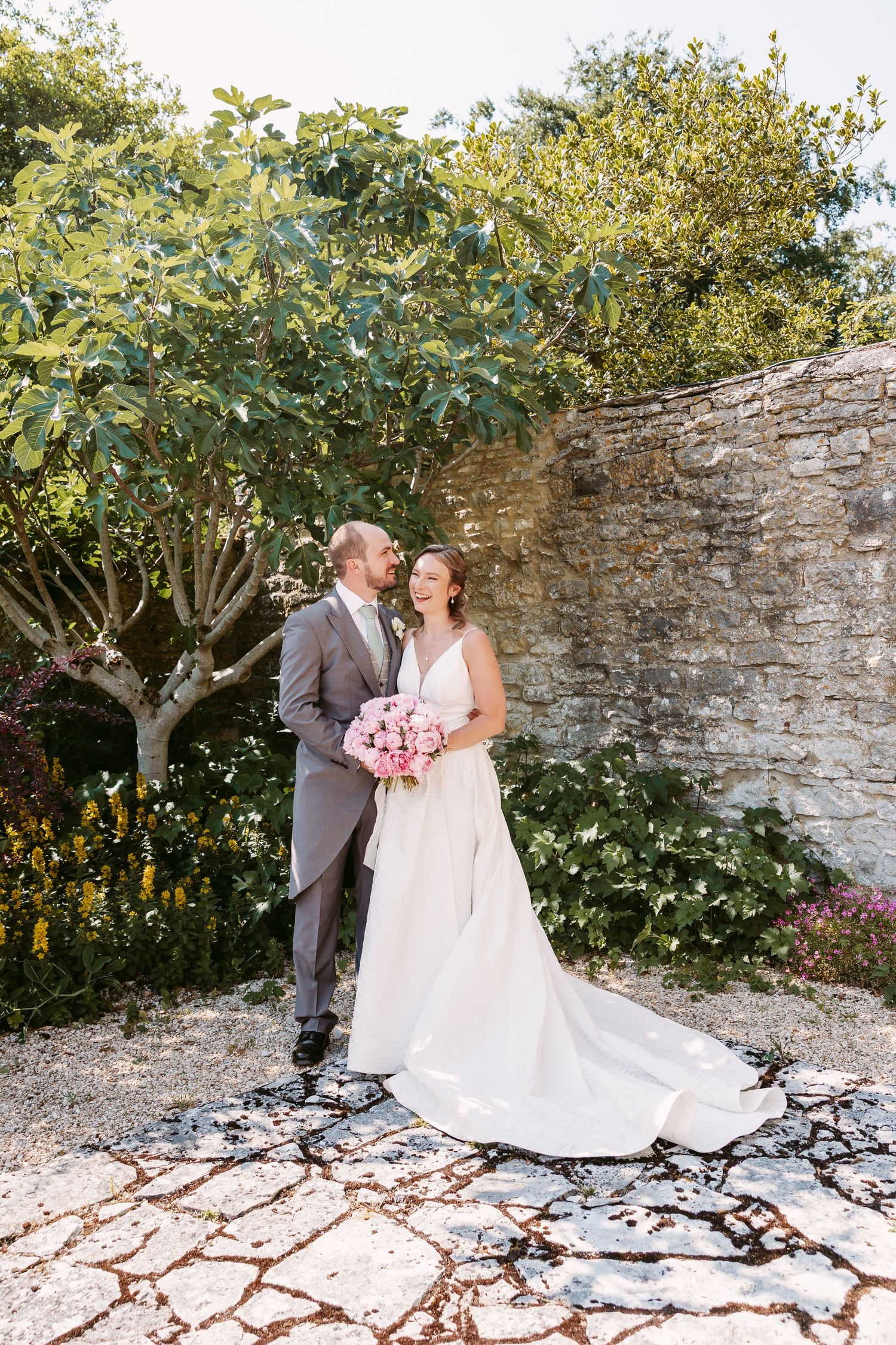 A romantic wedding couple poses together in front of a rustic stone wall and lush greenery in a garden setting.