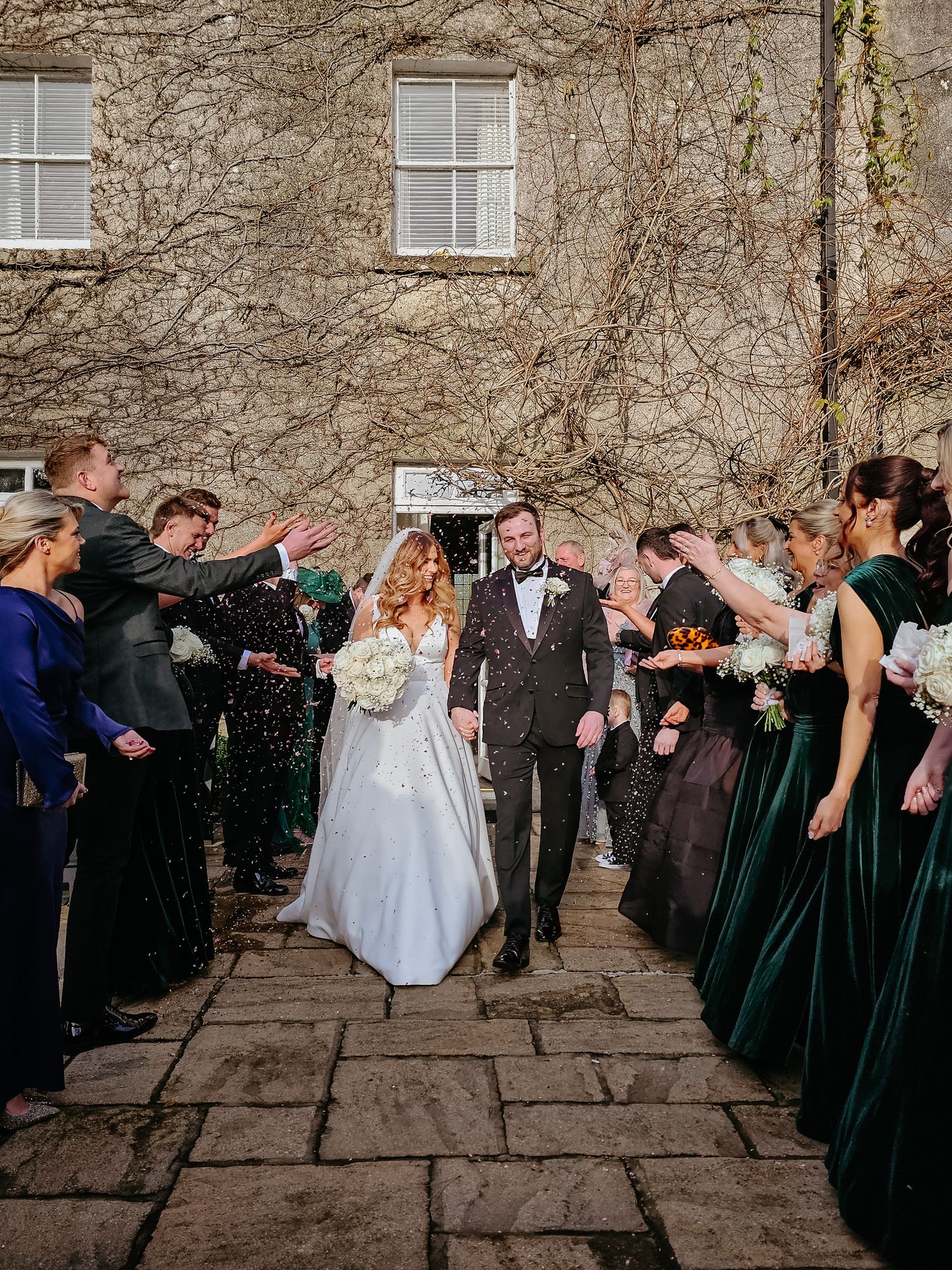 Wedding party performs a synchronized dip pose in front of a stone building during a celebration.