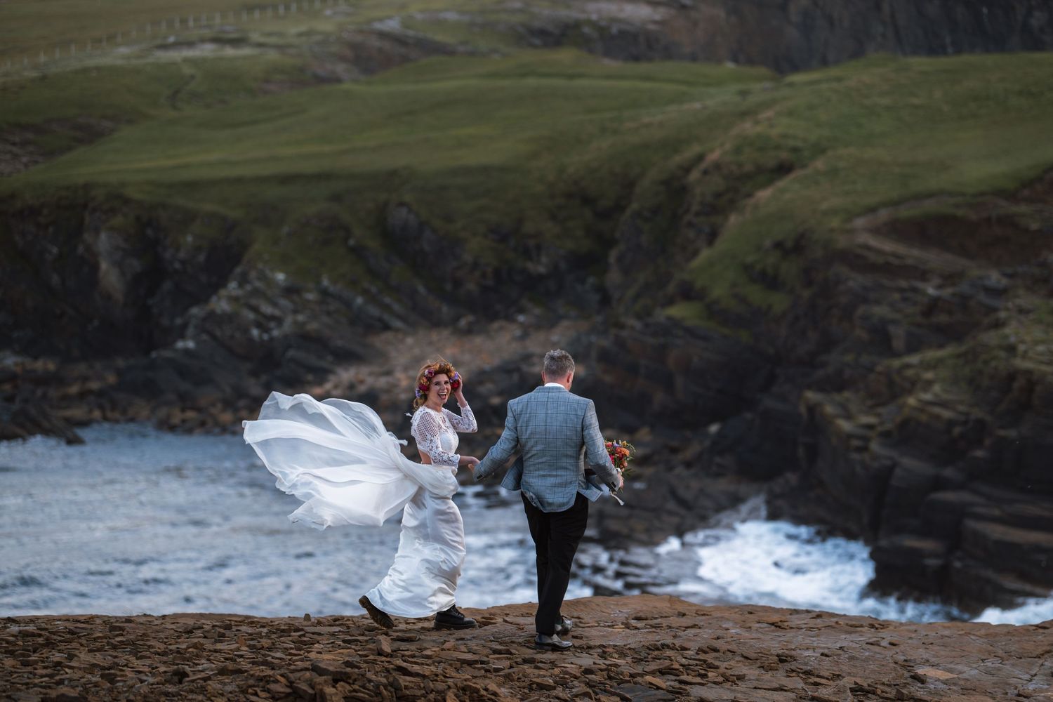A couple walks along dramatic coastal cliffs as a white wedding dress billows in the wind by the ocean.
