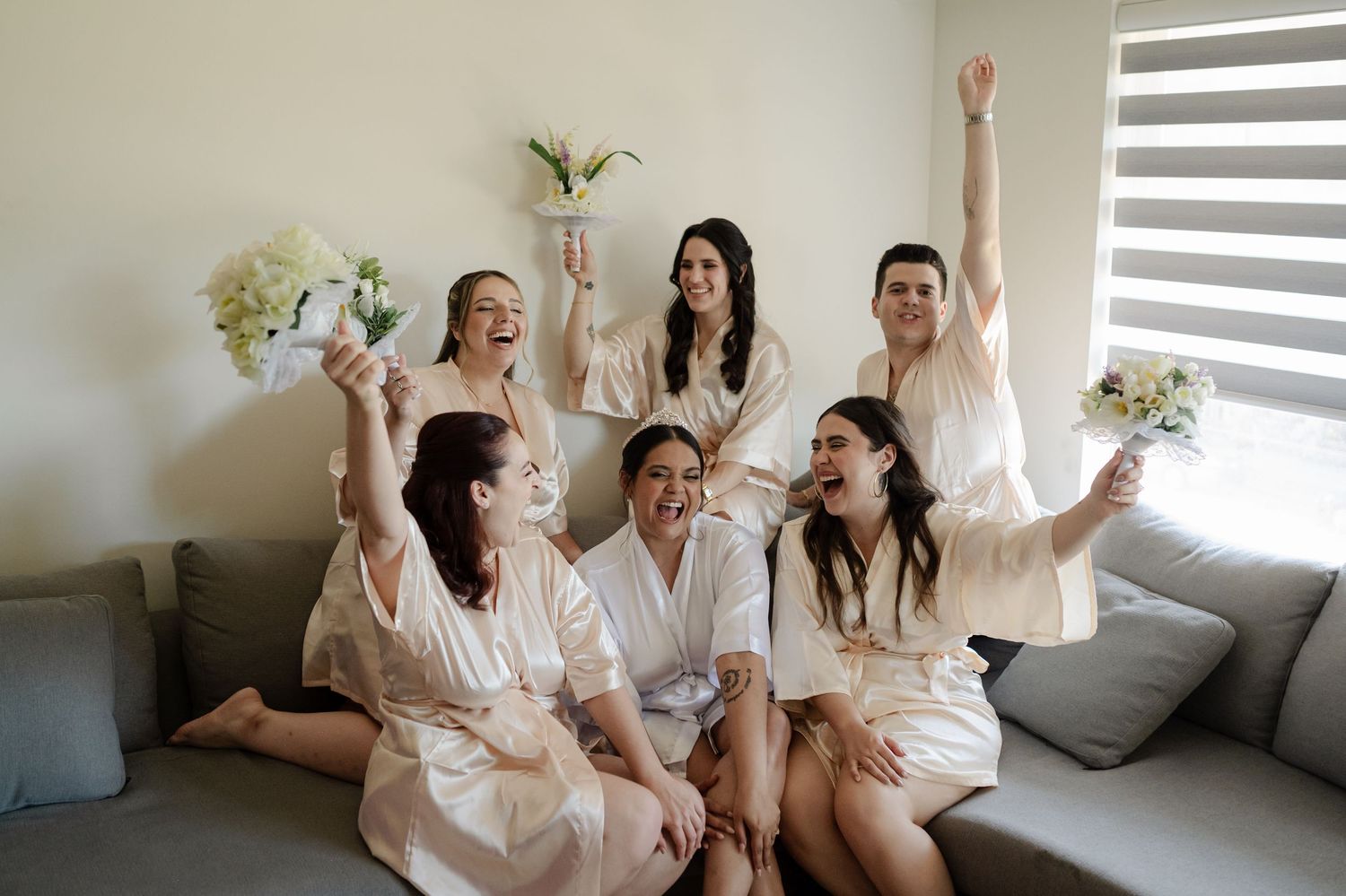 Group celebrating in matching silk robes with white floral bouquets on a gray couch.