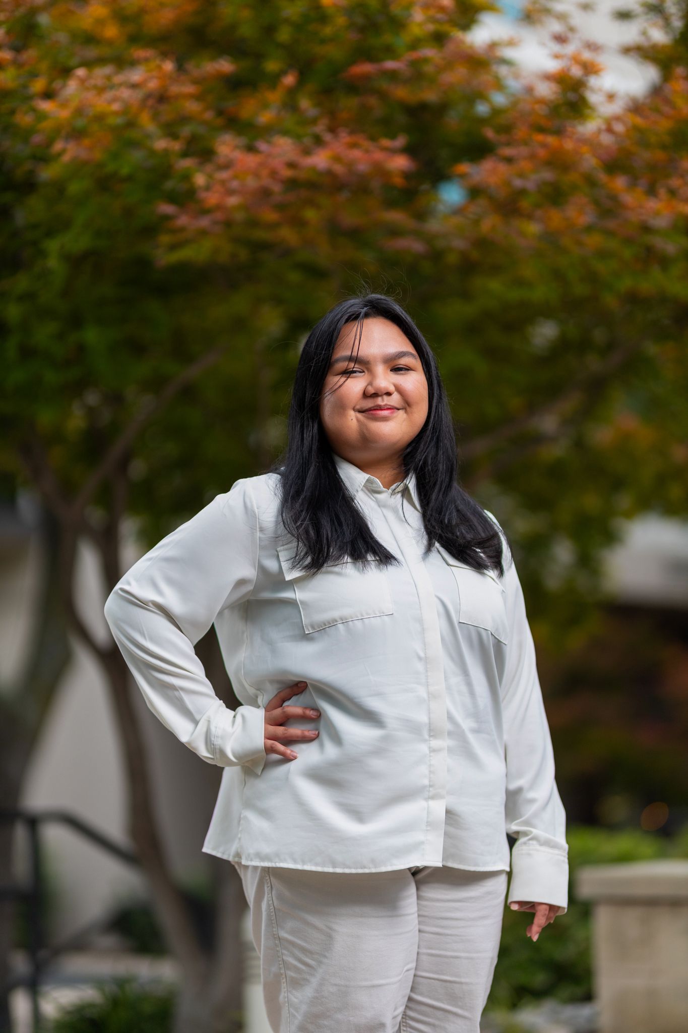 Person in all white outfit poses outdoors with autumn trees in the background.
