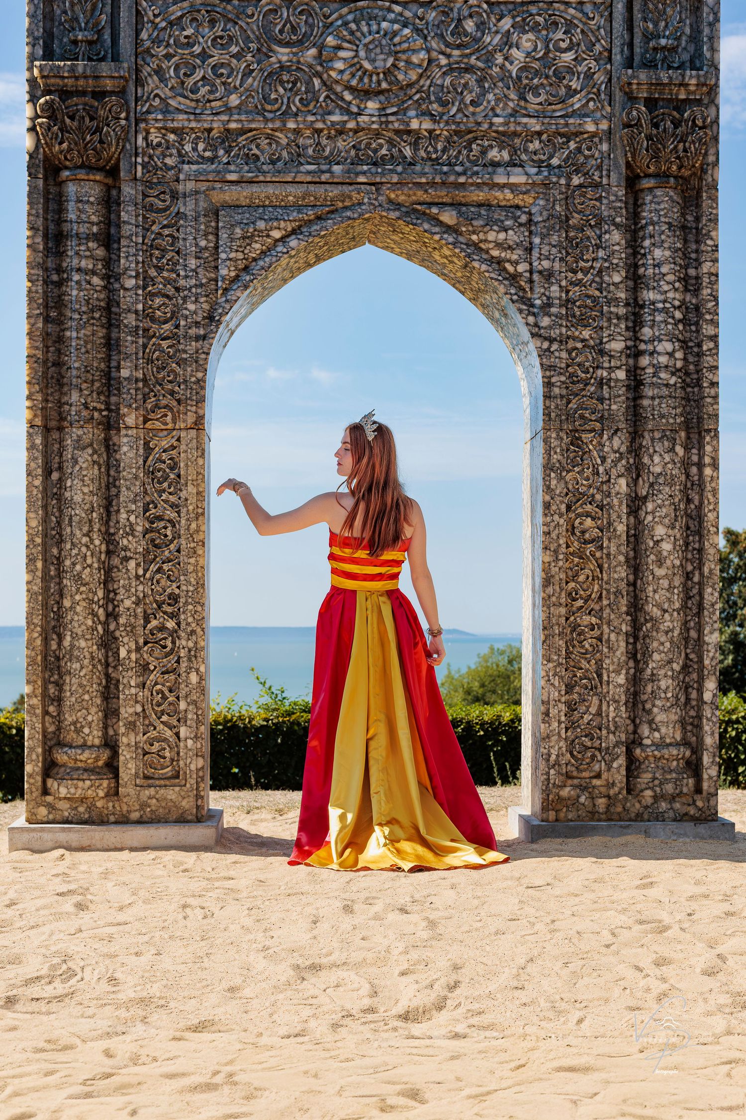 Red and yellow dress flows gracefully through ornate stone archways on sandy ground.