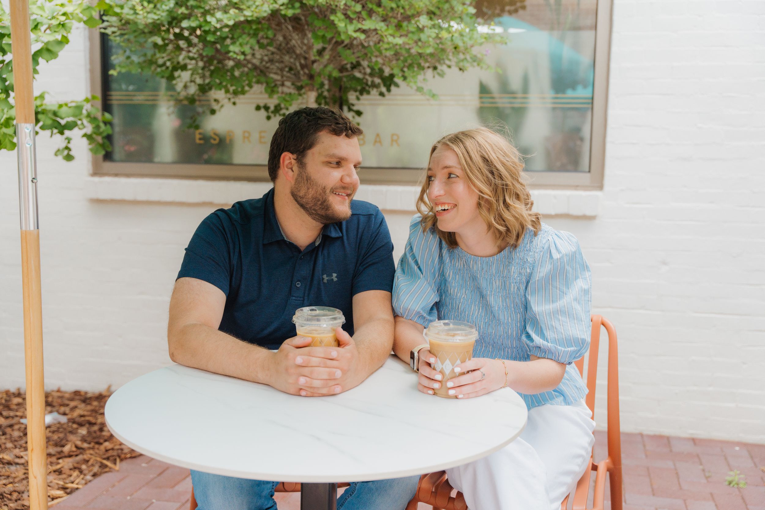A couple smiles at each other while sharing drinks at an outdoor cafe table, enjoying a sunny day together.
