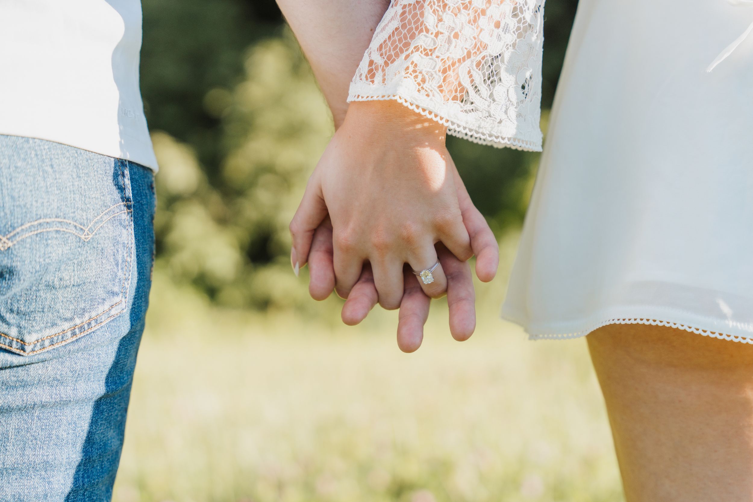 Close-up of a couple holding hands outdoors, woman wearing an engagement ring, with a green nature background.