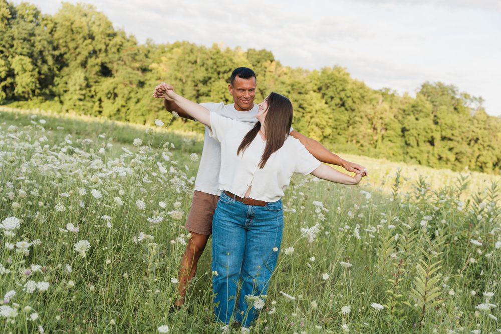 Couple dancing joyfully in a blooming wildflower meadow, wearing white shirts and jeans, surrounded by lush green trees.