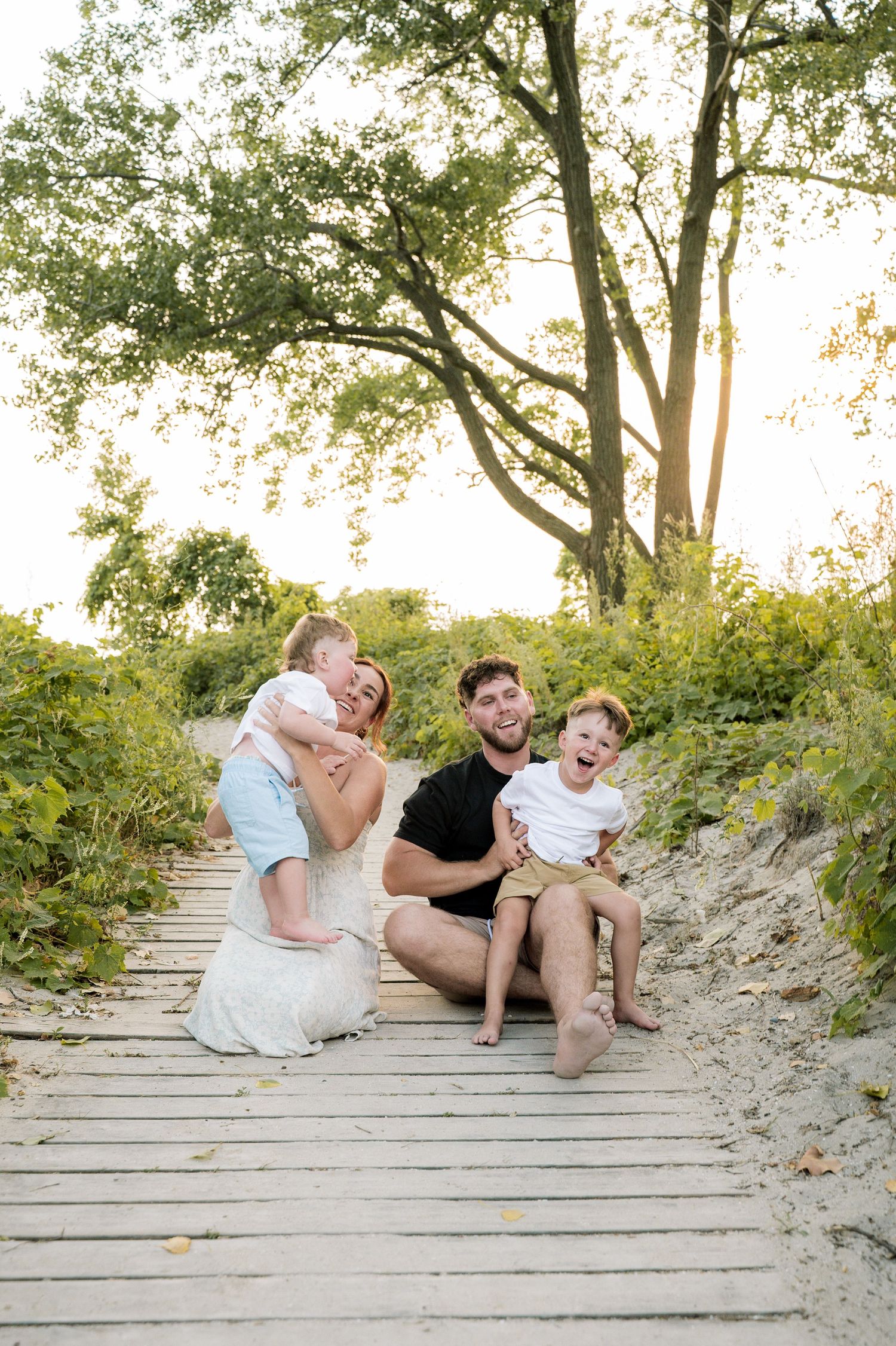 Family relaxing together on wooden boardwalk steps through sandy dunes at sunset.