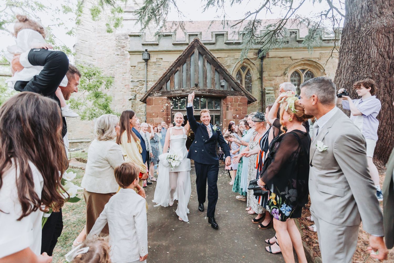 Wedding guests stand in a line outdoors tossing flower petals and confetti as newlyweds walk through the celebration.