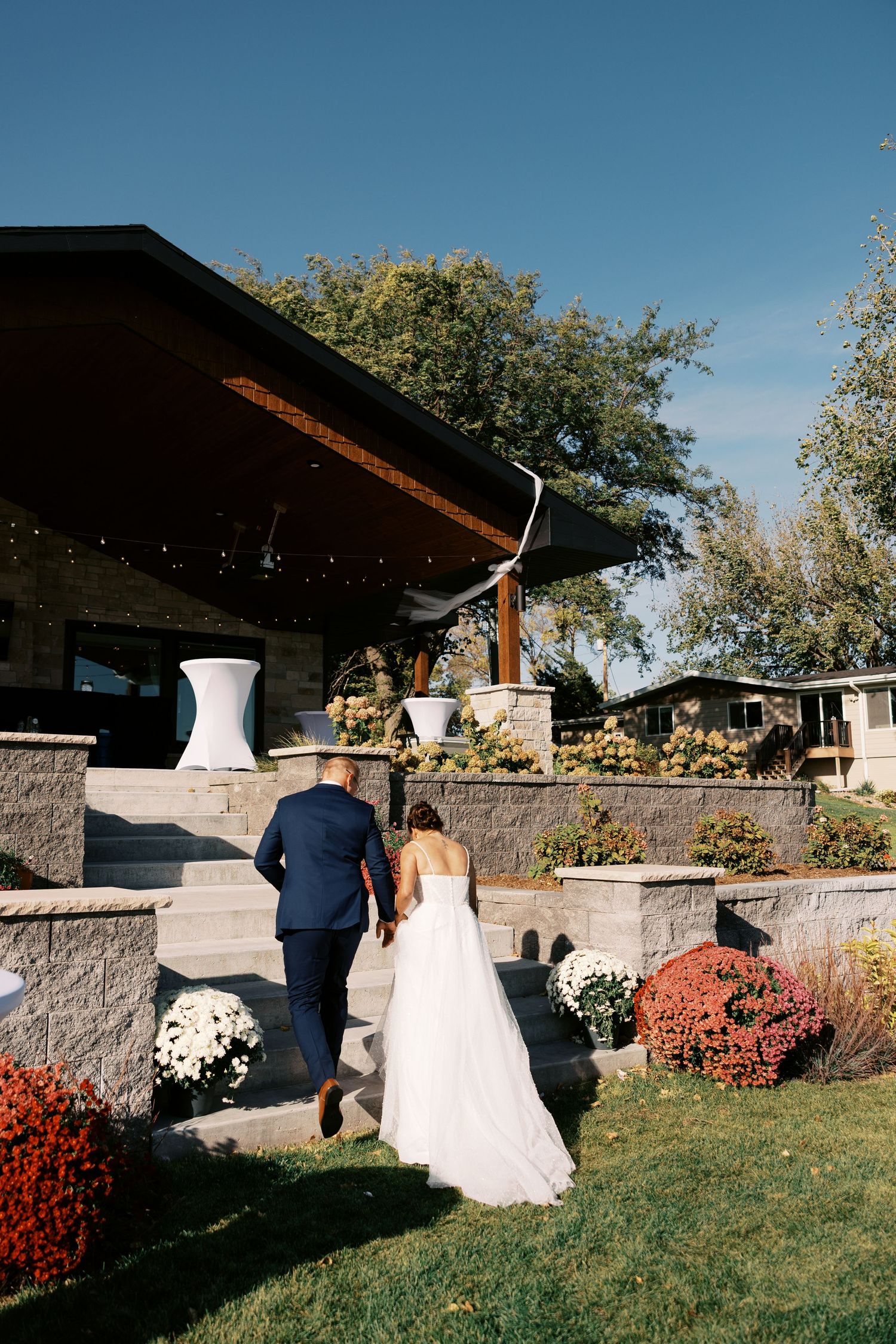 A couple in wedding attire walk together on a dirt path surrounded by trees on a sunny day.