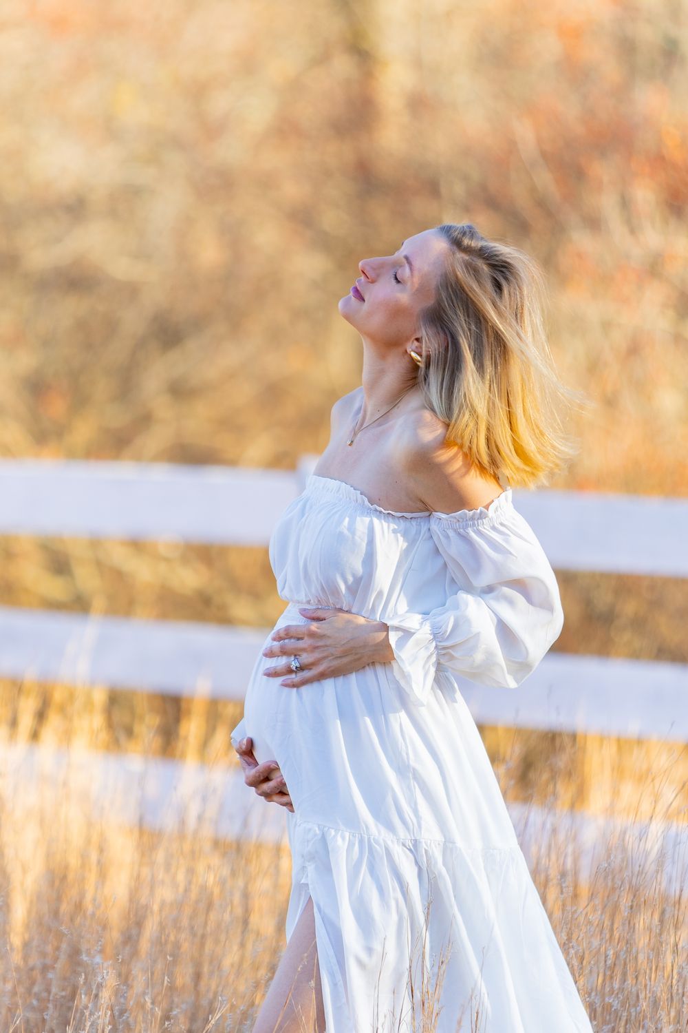 An expectant mother in a flowing white dress twirls joyfully in a golden sunlit field during sunset during maternity photoshoot.