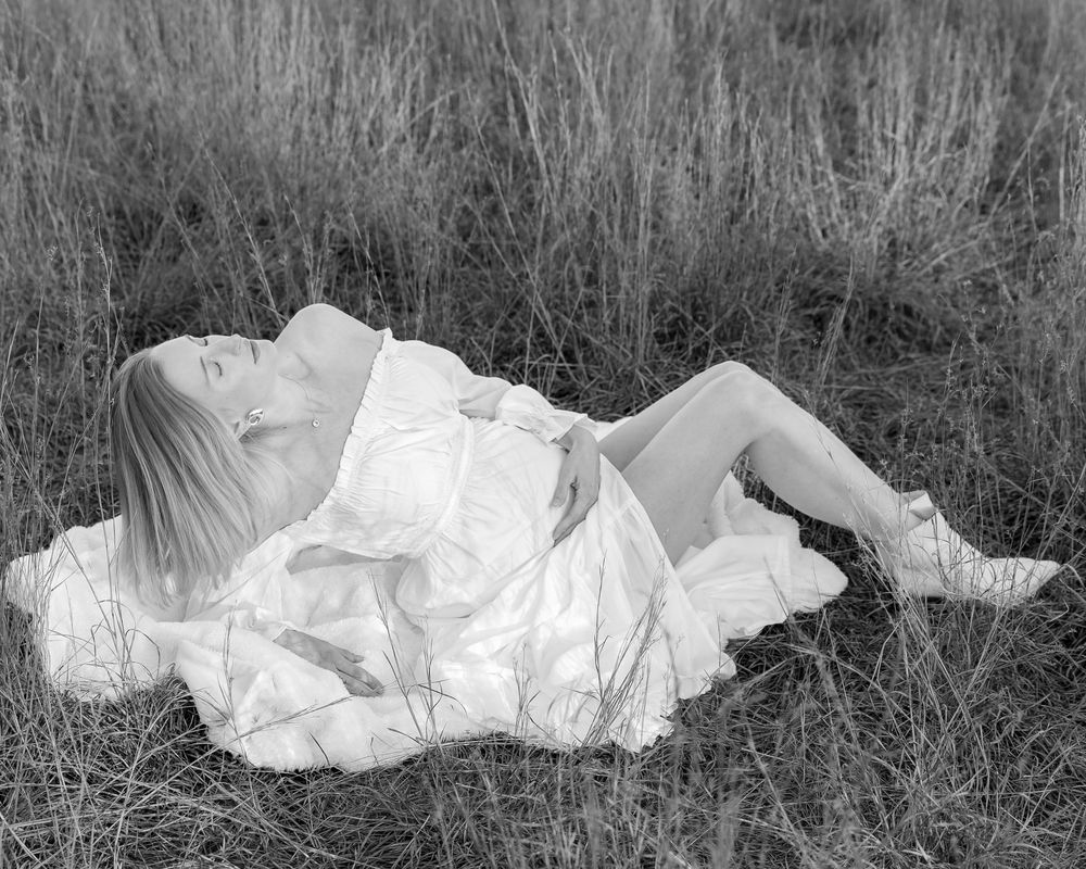 A dramatic black and white photo showing pregnant woman reclined in a grassy field cradling her belly during maternity photoshoot.