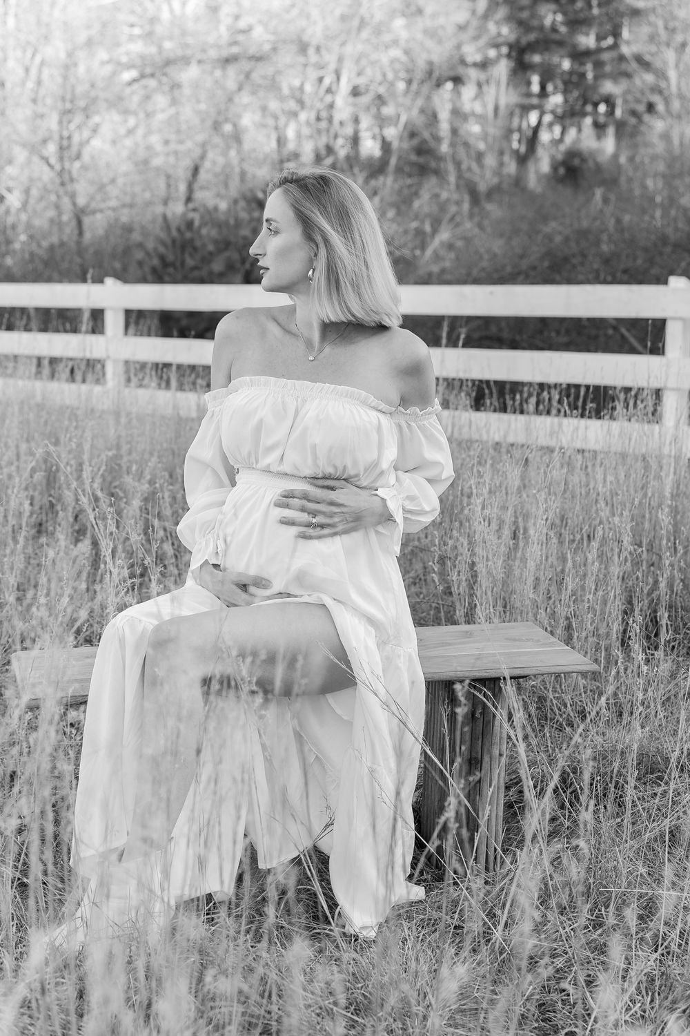 A black and white portrait of a person in a flowing white dress sitting on a bench in a rustic field with a fence during a maternity photos.
