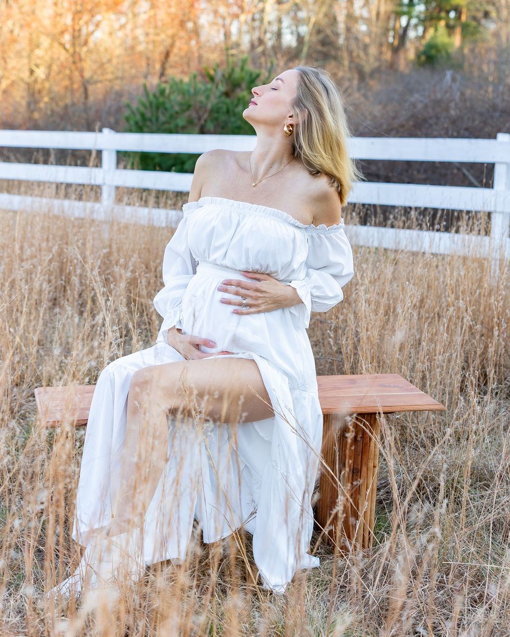 Ethereal maternity photoshoot in a golden field with white dress and white fence at sunset.