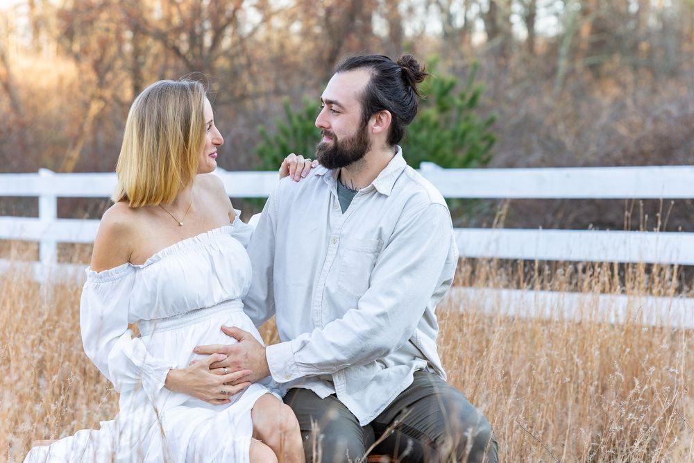 A couple in white outfits share romantic moments in a golden field during sunset with a white fence in background during maternity photos.