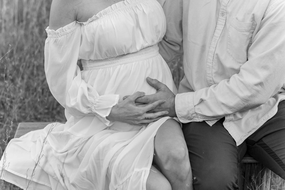 A black and white photograph shows a couple in embracing a pregnant belly in a tall grass field in Metrowest area of Boston, MA.