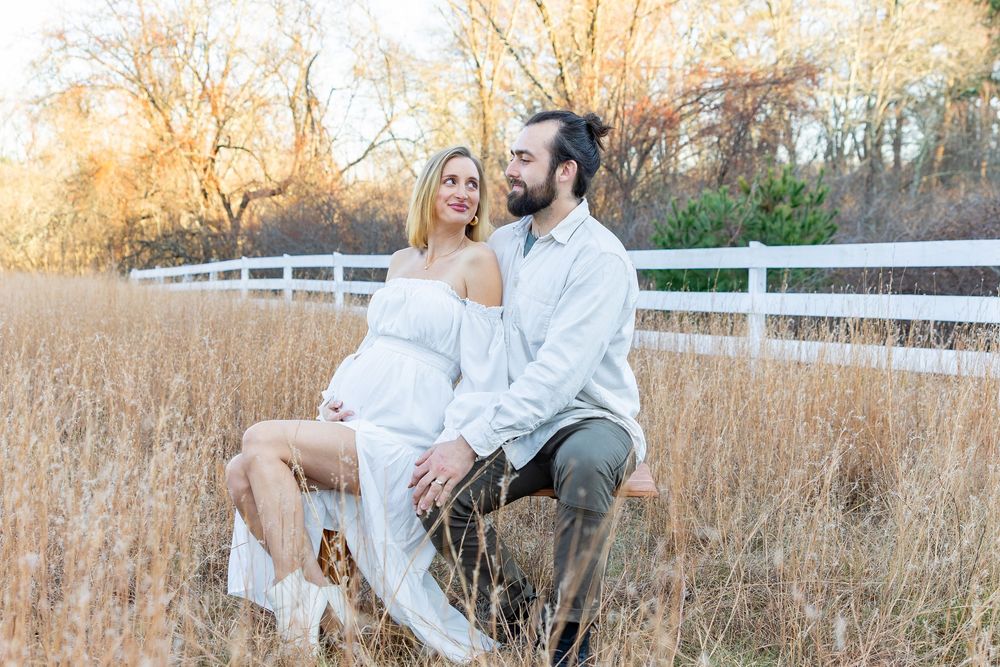 A couple in white outfits poses together in a rustic outdoor setting with tall grass during golden hour at a maternity photo session .