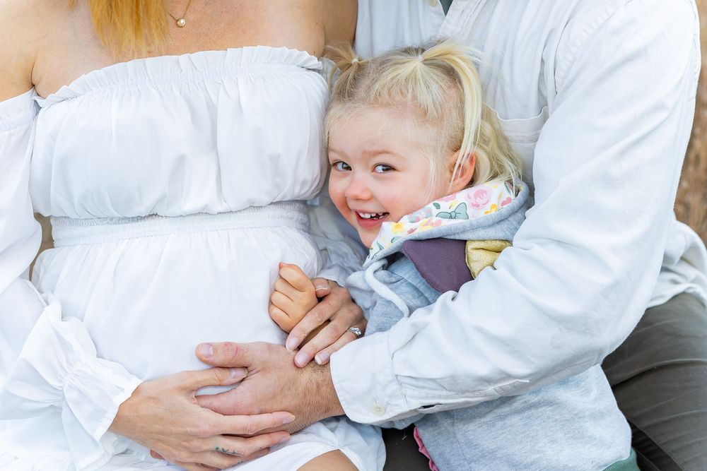 A tender moment showing parents in white shirts holding and a toddler cradling a pregnant belly during a maternity photo session.