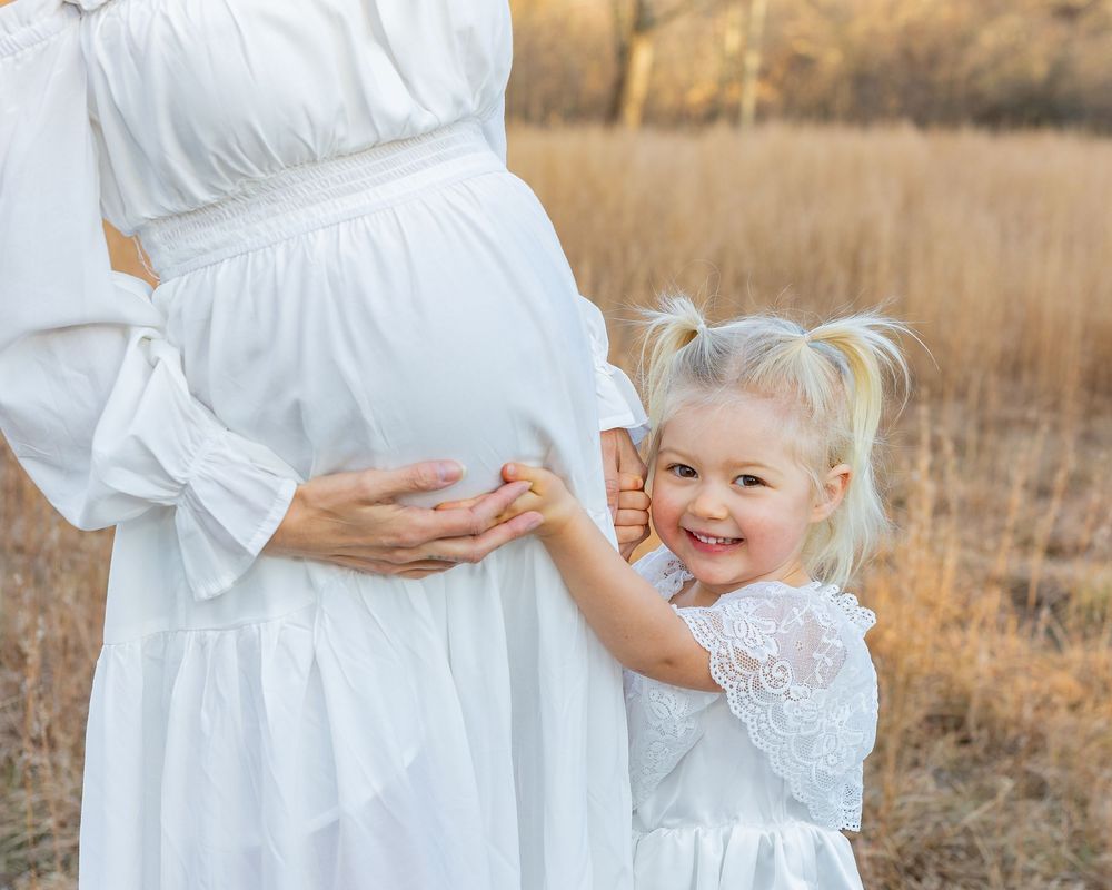 A pregnant mother and toddler in white dresses stands together in a golden field during sunset in Metrowest of Boston- Sudbury.