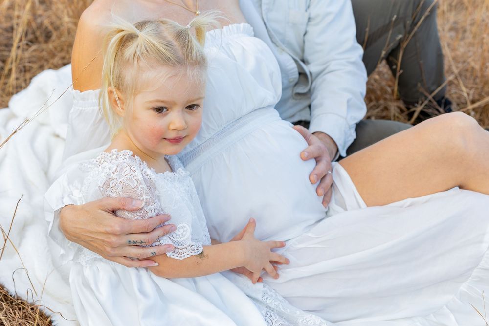 A family in white flowing dresses relax together in a grassy outdoor setting during a maternity photo shoot in Sudbury, MA