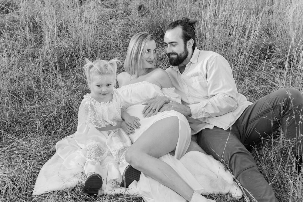 A black and white photo shows a family relaxing together in a grassy outdoor setting during a maternity photo shoot in Metrowest of Boston.