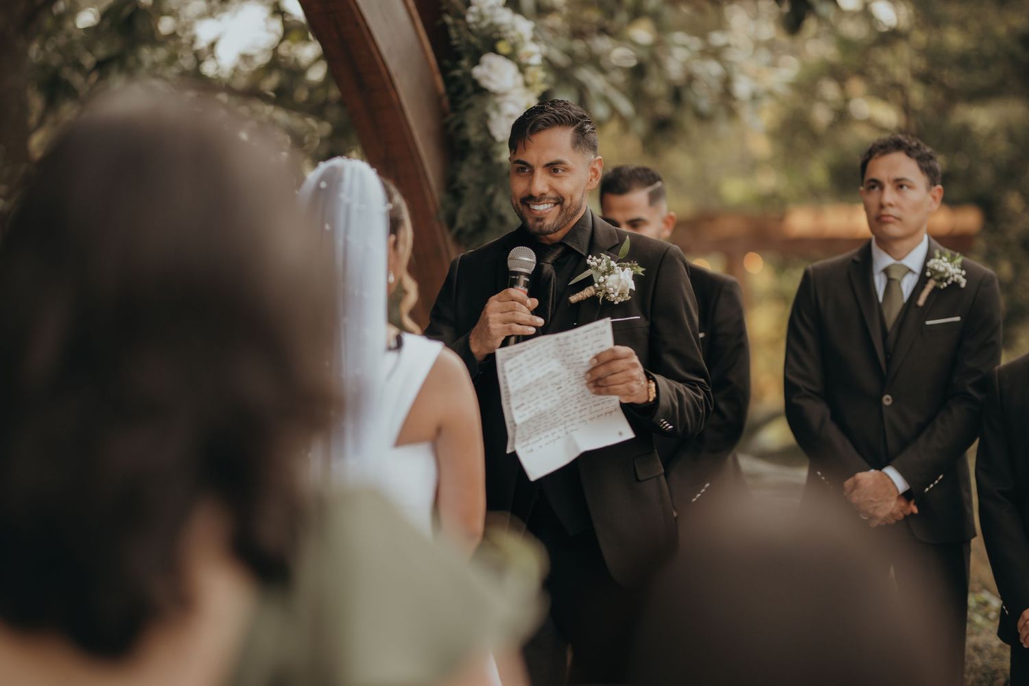 Wedding guests watch with joy during an outdoor ceremony with trees in the background.