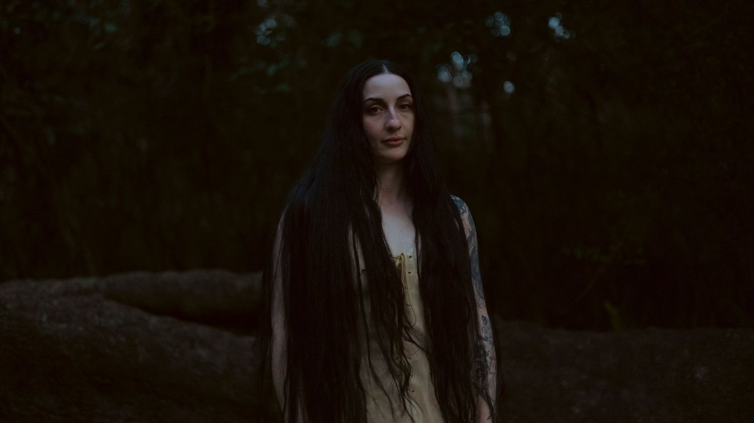 A woman with long dark hair poses in a forest setting wearing black clothing in a moody atmospheric photoshoot.