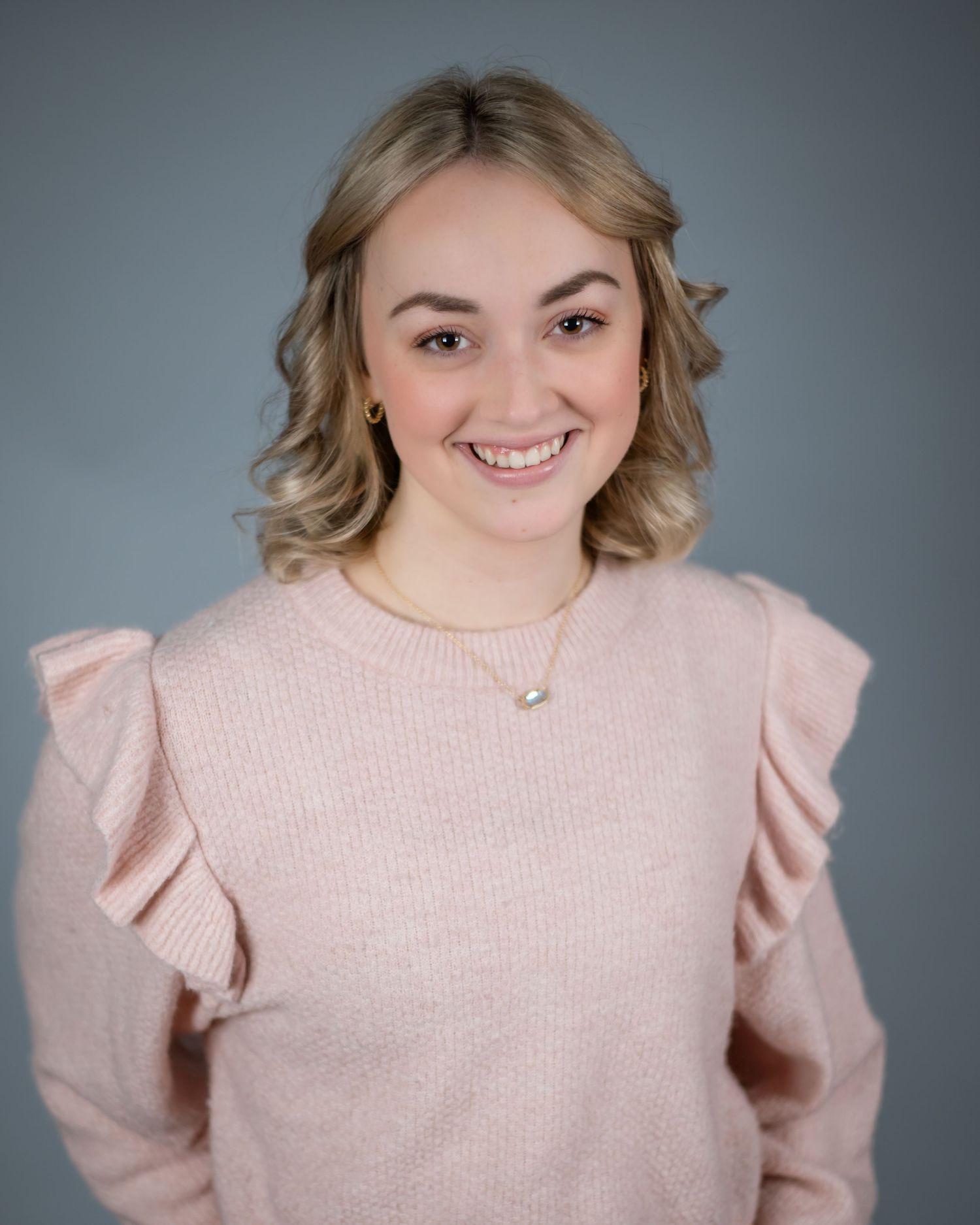 Professional headshot of someone wearing a soft pink sweater with ruffled shoulders against a gray background.