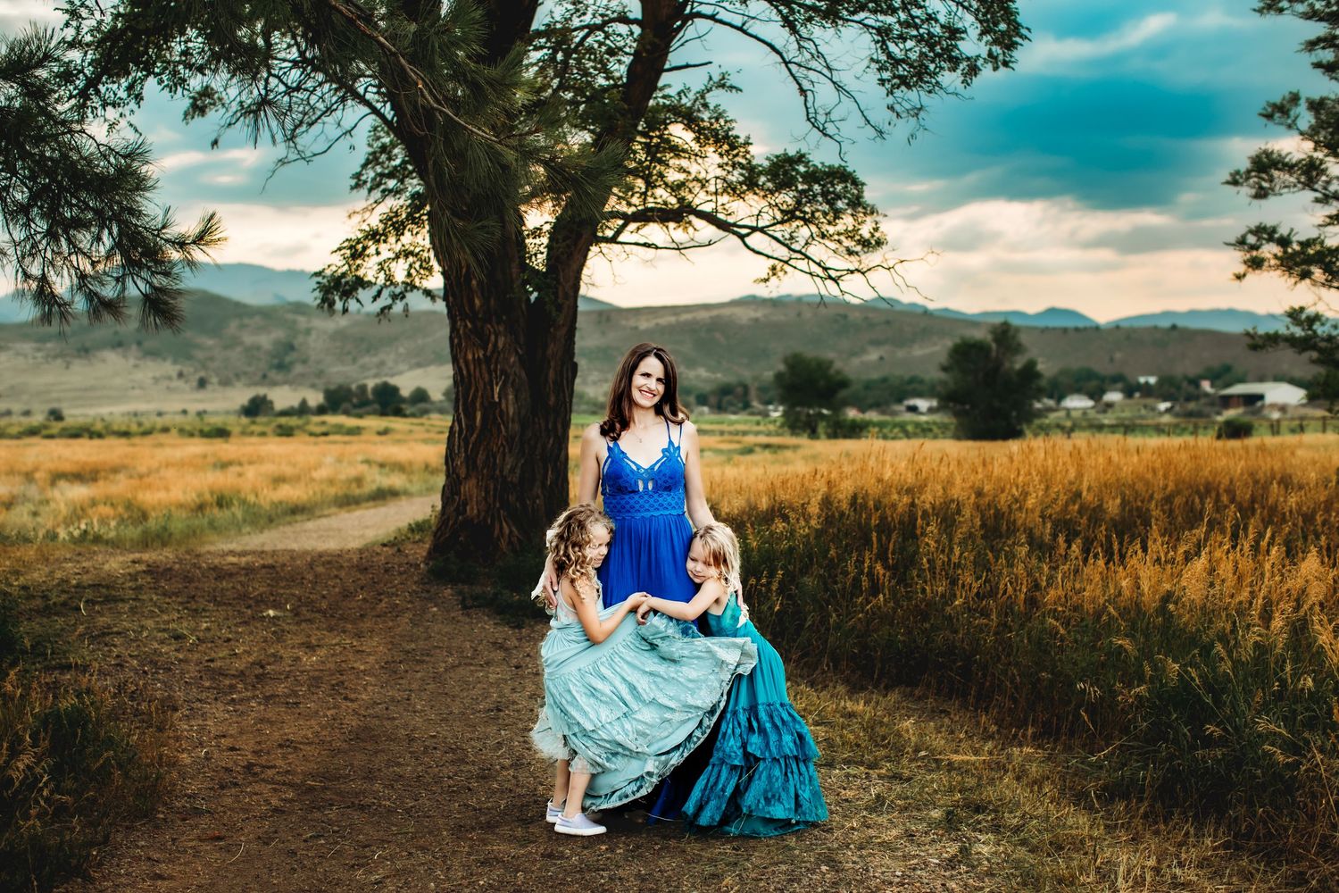 A person in a flowing blue dress poses in a golden field at sunset with mountains in the background.