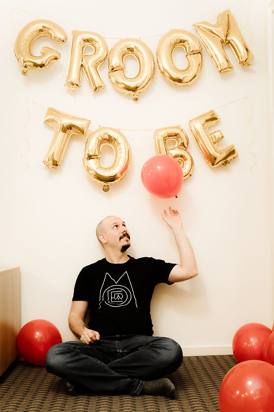 A person in black clothing sits on the floor with gold letter balloons and red balloons against a white wall.