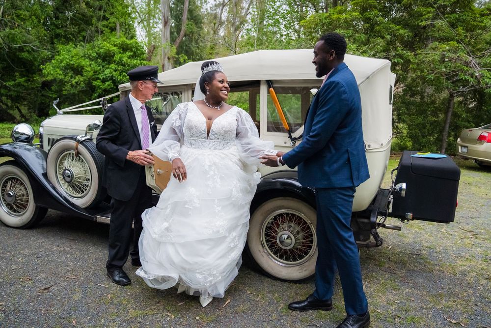 A wedding party poses next to a vintage white car during an outdoor celebration.