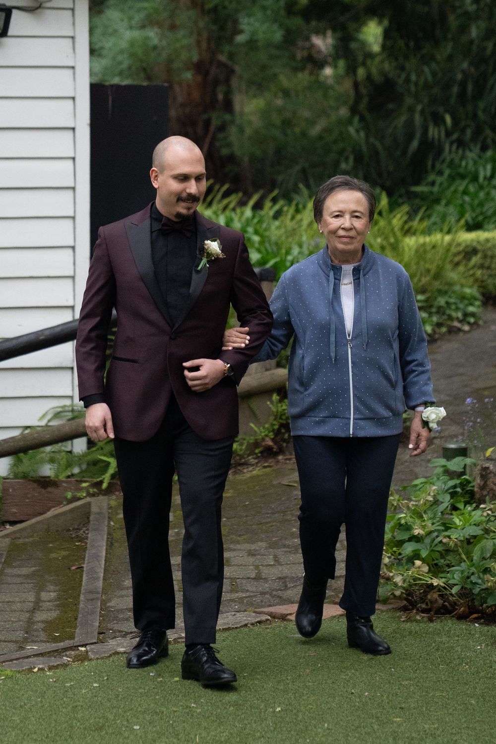 Two people walking together outdoors, one in a burgundy suit jacket and one in a blue fleece jacket, near garden foliage.