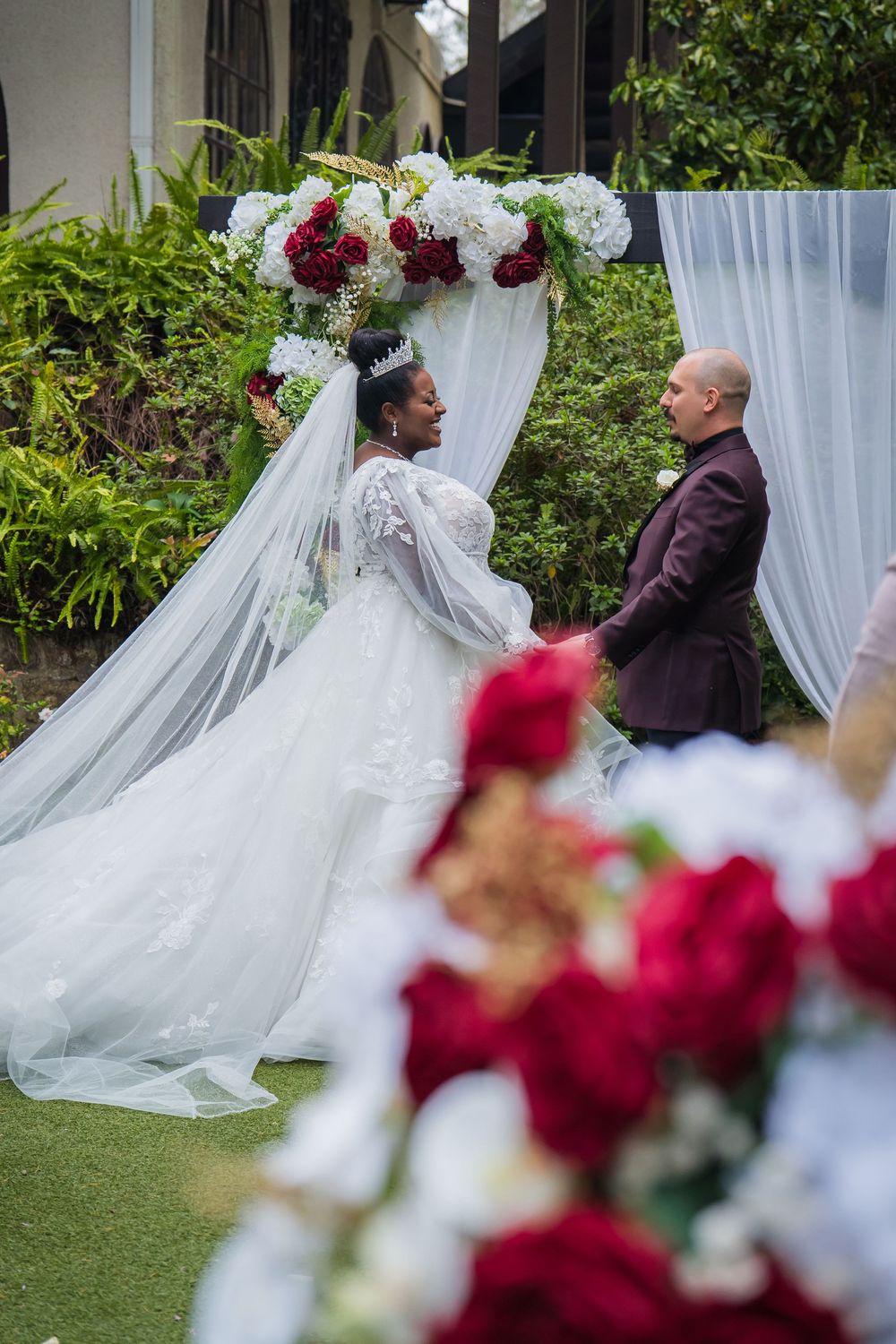 A wedding ceremony scene with flowing white tulle drapes and deep red floral arrangements in an outdoor garden setting.