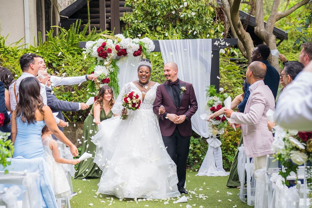 Outdoor garden wedding ceremony with guests seated in white chairs surrounded by lush greenery and floral arrangements.