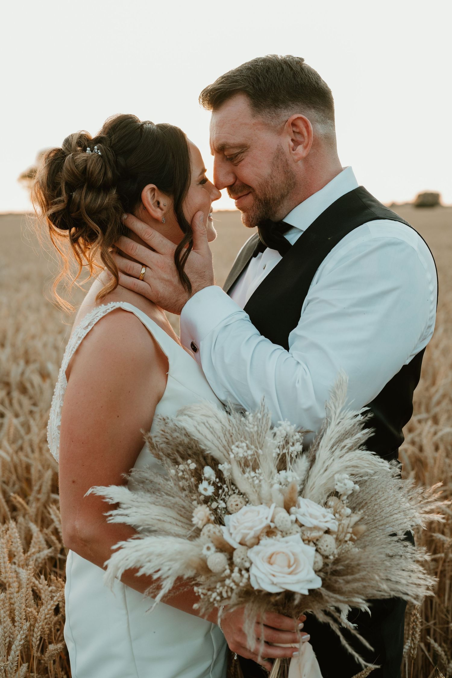 A romantic wedding couple embraces in a golden field at sunset holding a pampas grass and rose bouquet.