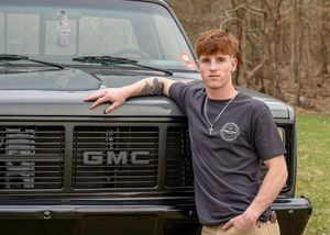 Casual pose leaning against gray GMC truck outdoors.