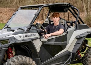 Driver seated in silver Turbo off-road side by side in wooded setting.