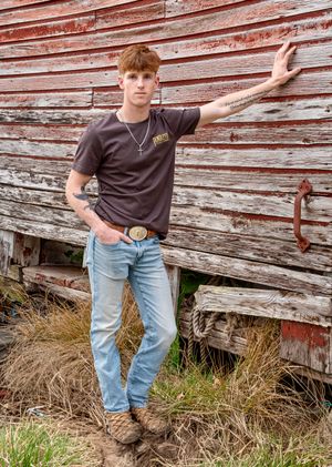 Casual pose against weathered red barn siding wearing brown t-shirt and jeans.