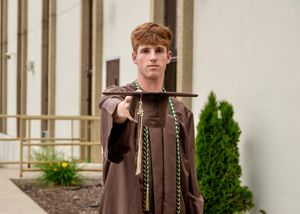 Graduate holding cap in brown graduation gown outdoors.