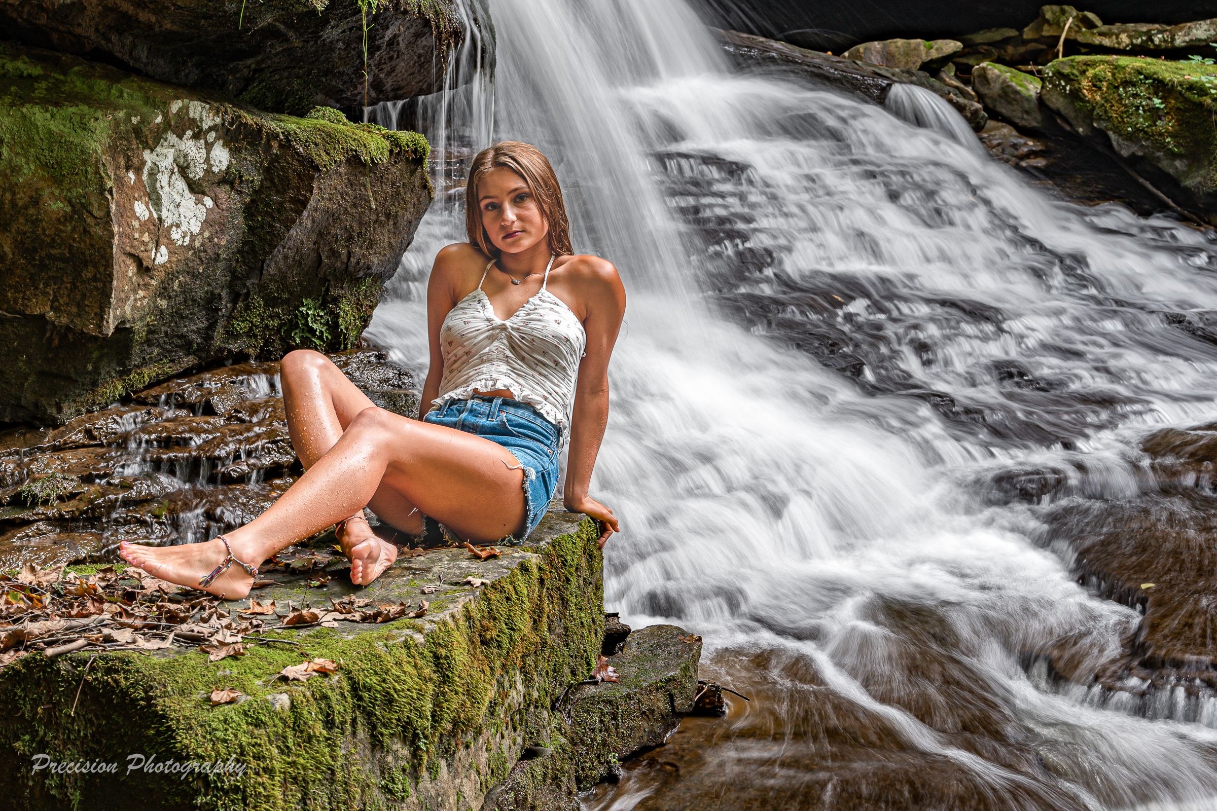 A cascading waterfall flows over mossy rocks in a lush forest setting while someone relaxes near the water's edge.