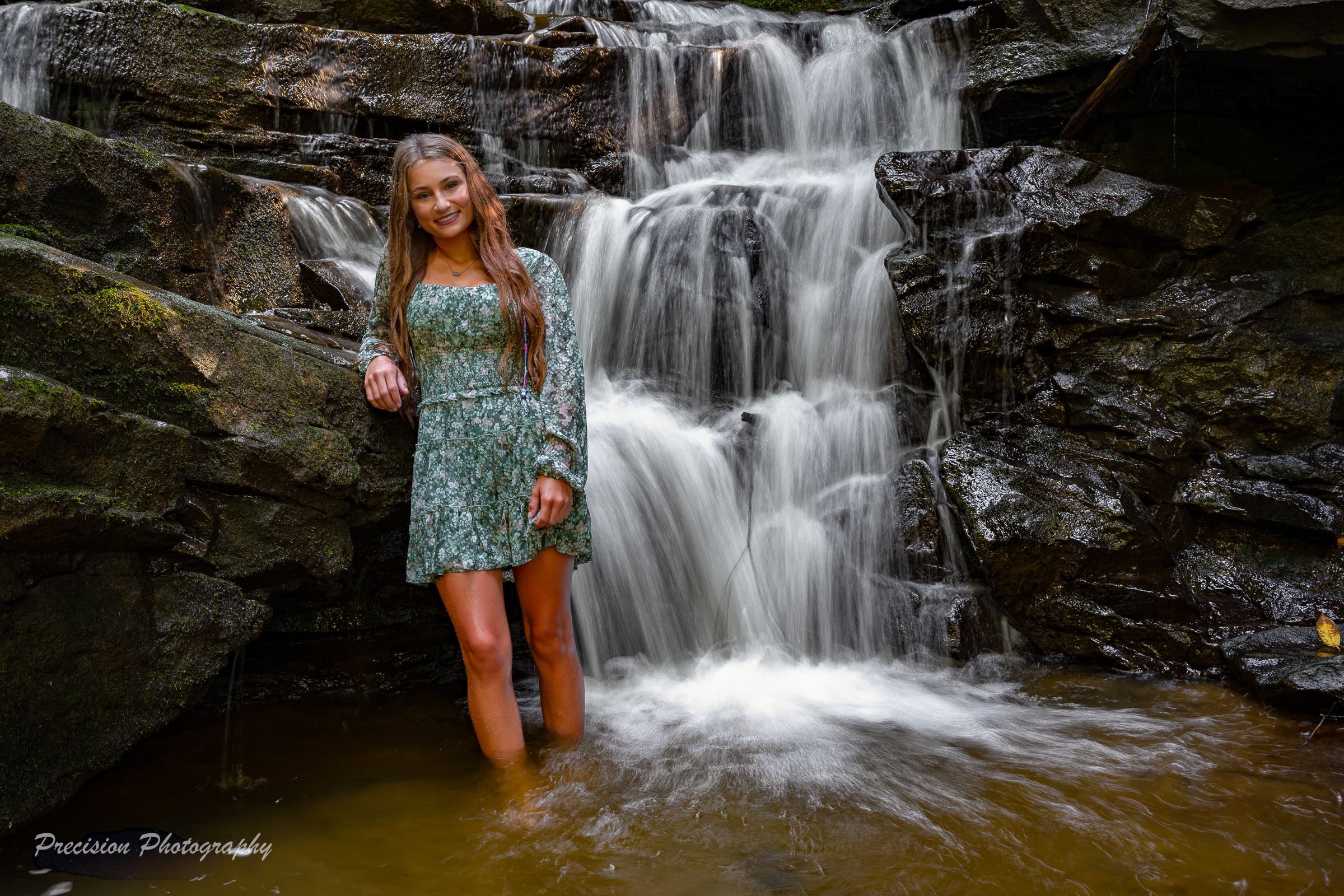 A person in a floral dress stands in front of a cascading waterfall on dark rocky cliffs.