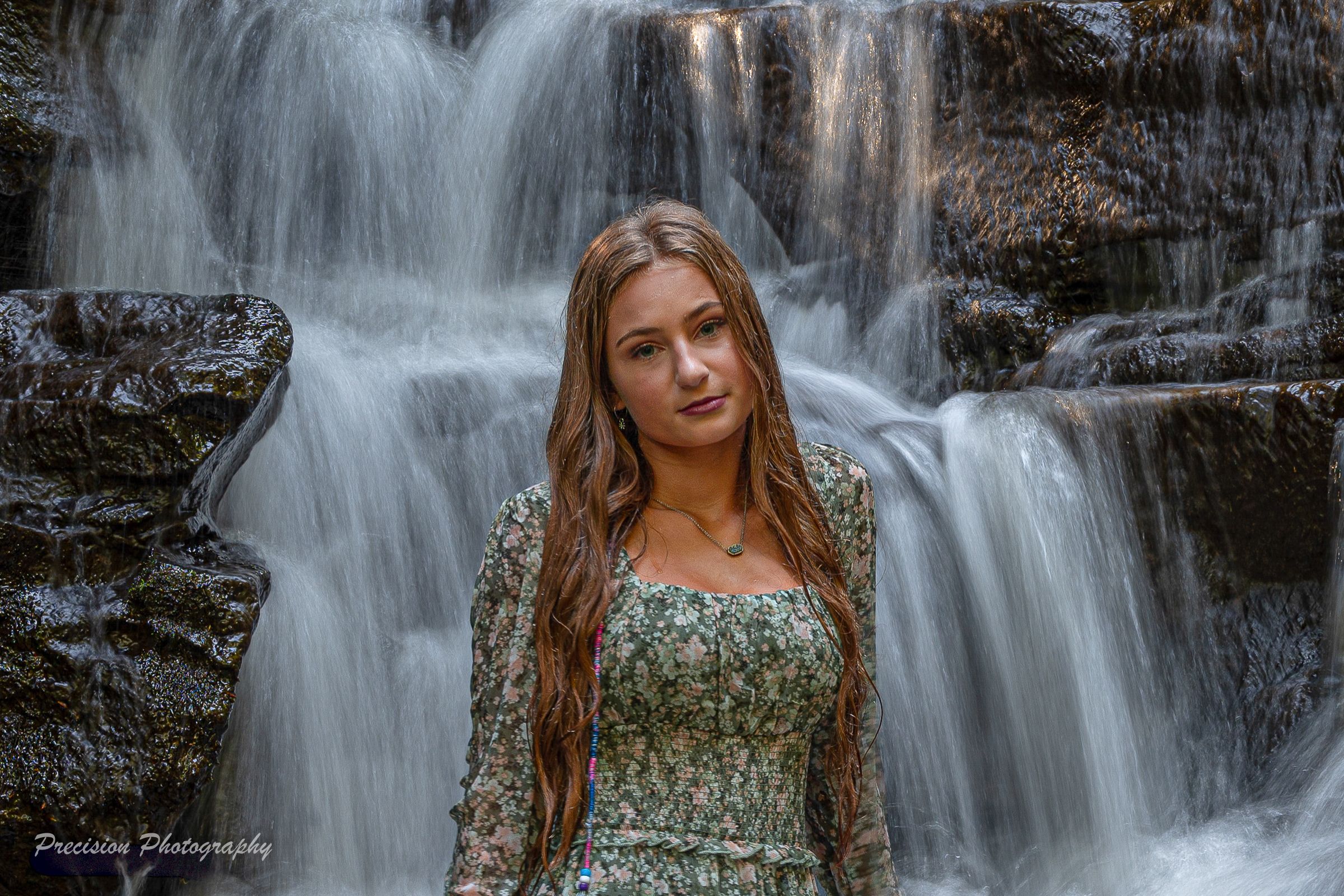 A person in a floral dress poses near a cascading waterfall in a scenic natural setting.