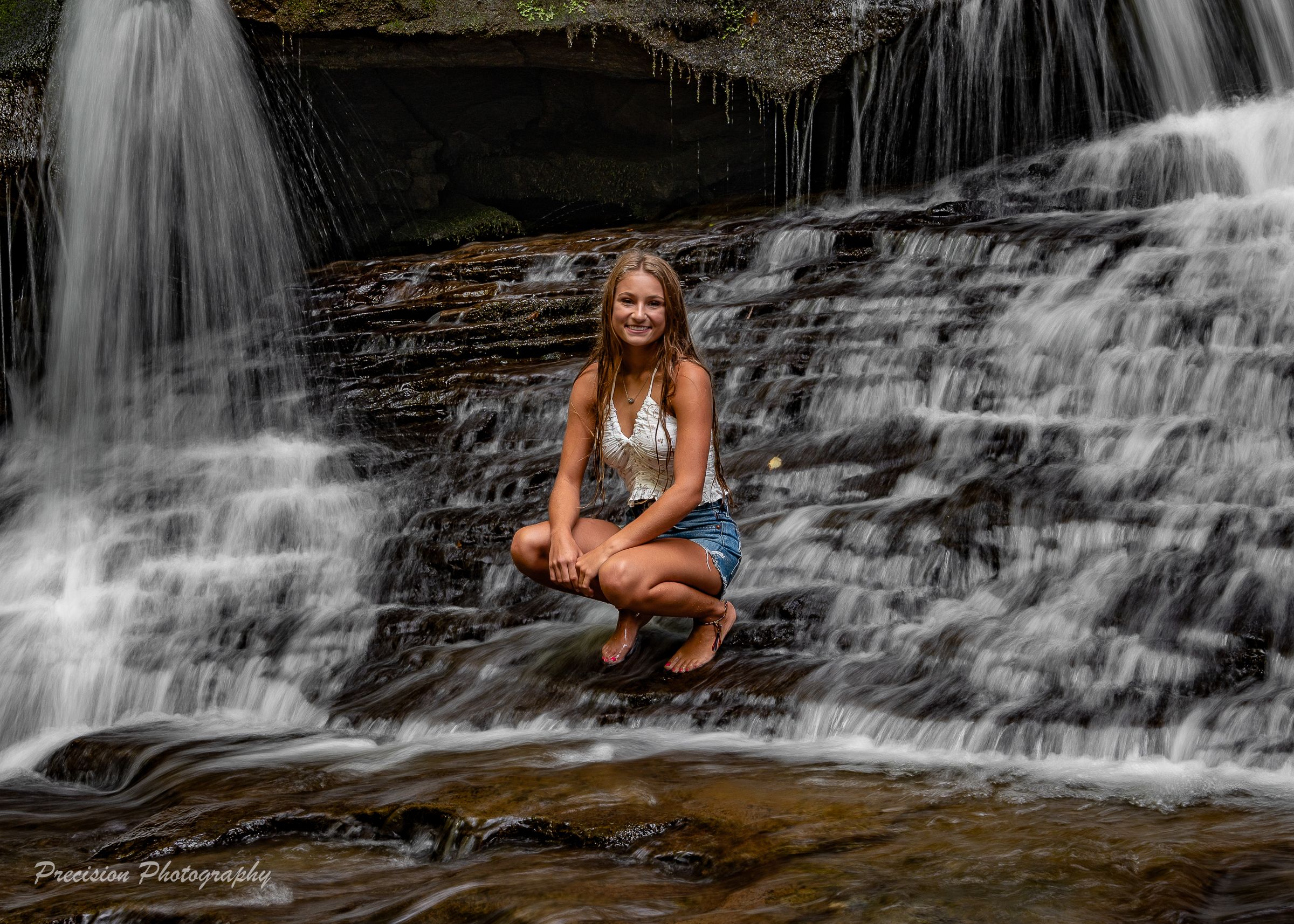 Someone poses on rocks in front of a cascading waterfall wearing summer attire and creating an outdoor portrait.
