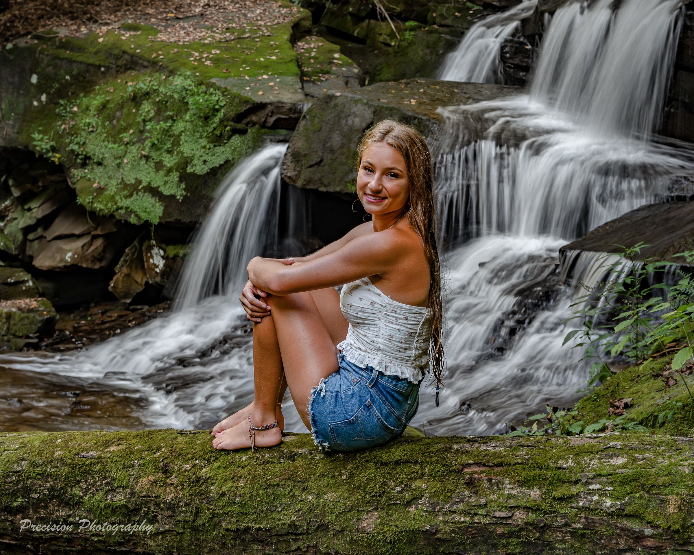 Cascading waterfall in lush forest setting with someone sitting on mossy rocks by the flowing water.