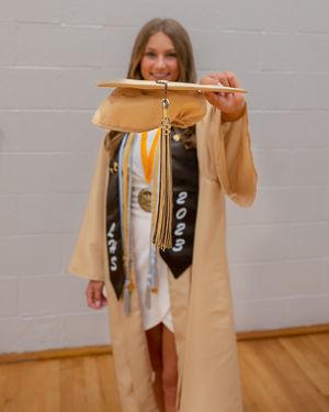 Graduate in tan cap and gown holds up mortarboard while wearing decorated stole and white dress for graduation photos.