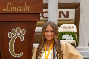 Graduate stands in front of wooden wall with Lincoln signage wearing yellow stole and graduation cap and gown.