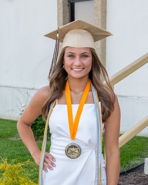 Graduate wearing white dress and gold medal poses outdoors in cap and gown for graduation portrait.