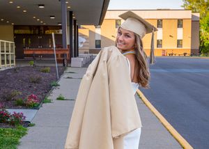 Graduate in white cap and gown poses outdoors in front of building with beige coat on sunny day.