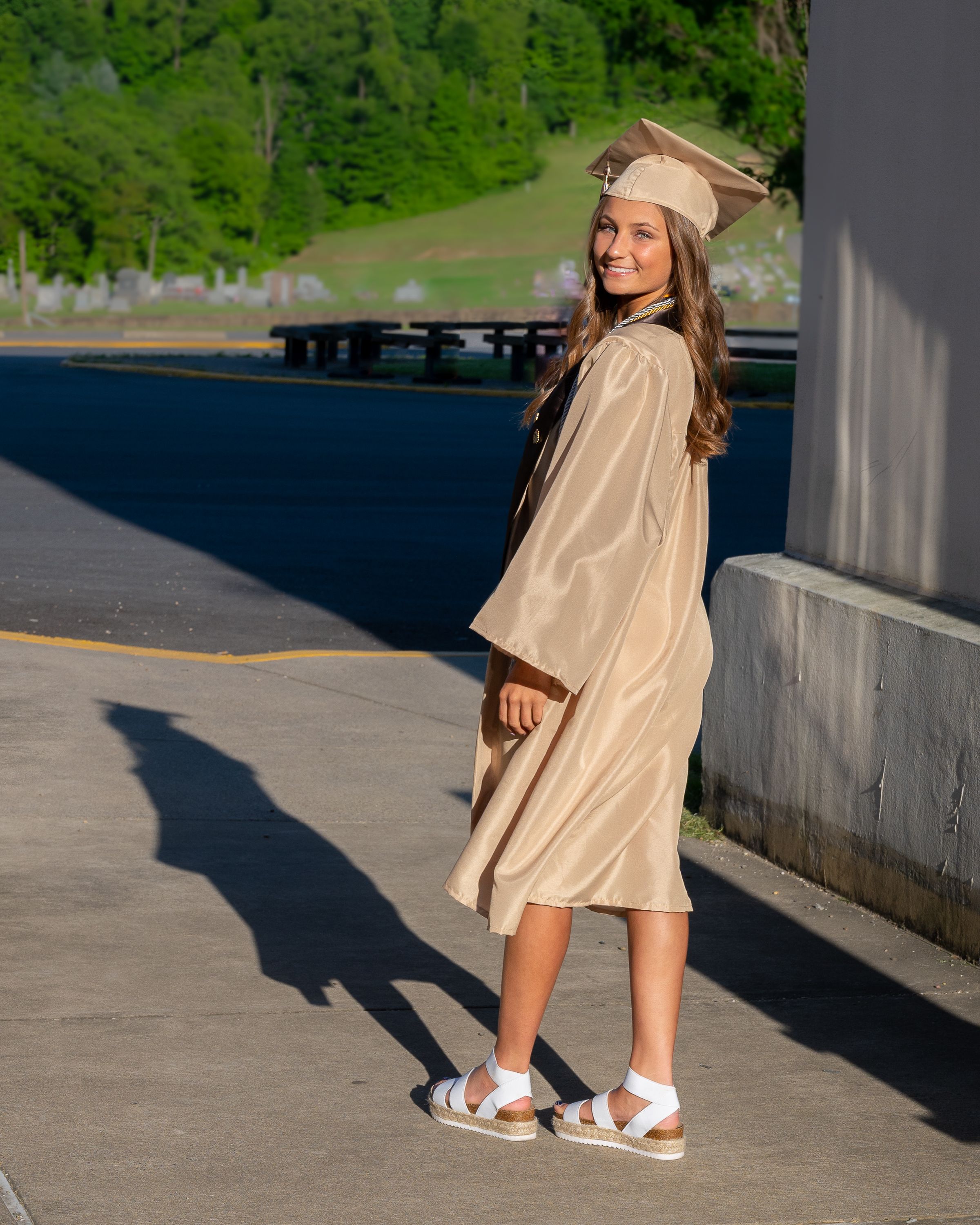Graduate in beige cap and gown poses on concrete walkway with green hills in background.
