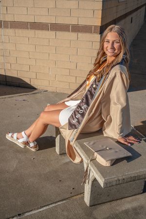 Graduate in beige coat and white dress sits on concrete bench outside brick building in sunlight.