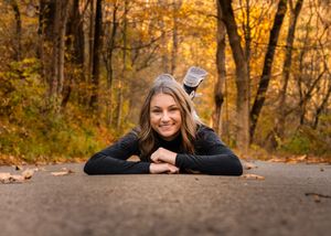 A person lies on a paved path surrounded by vibrant autumn foliage in a forest setting during golden hour.