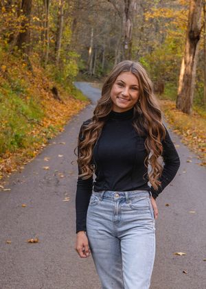 A person in a black turtleneck and light jeans walks along a tree-lined autumn path covered in fallen leaves.